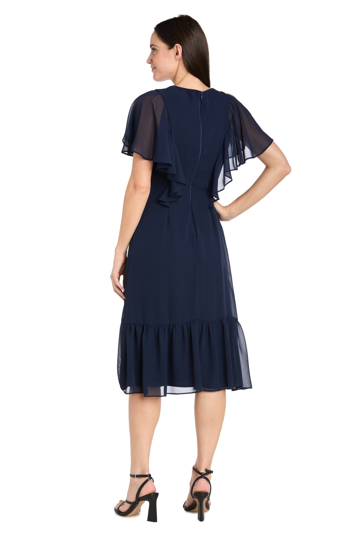 Woman models the R&M Richards 4054P Petite Navy Blue Midi Party Dress with flutter sleeves and ruffled hem, posing with her back to the camera and hand on hip, paired with black heeled sandals.